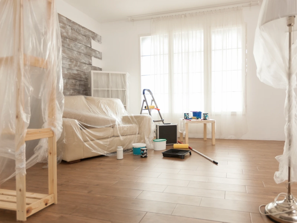 Sofa zabezpieczona folią, wiadra z farbą, wałek i drabina w pokoju podczas usługi Malowanie