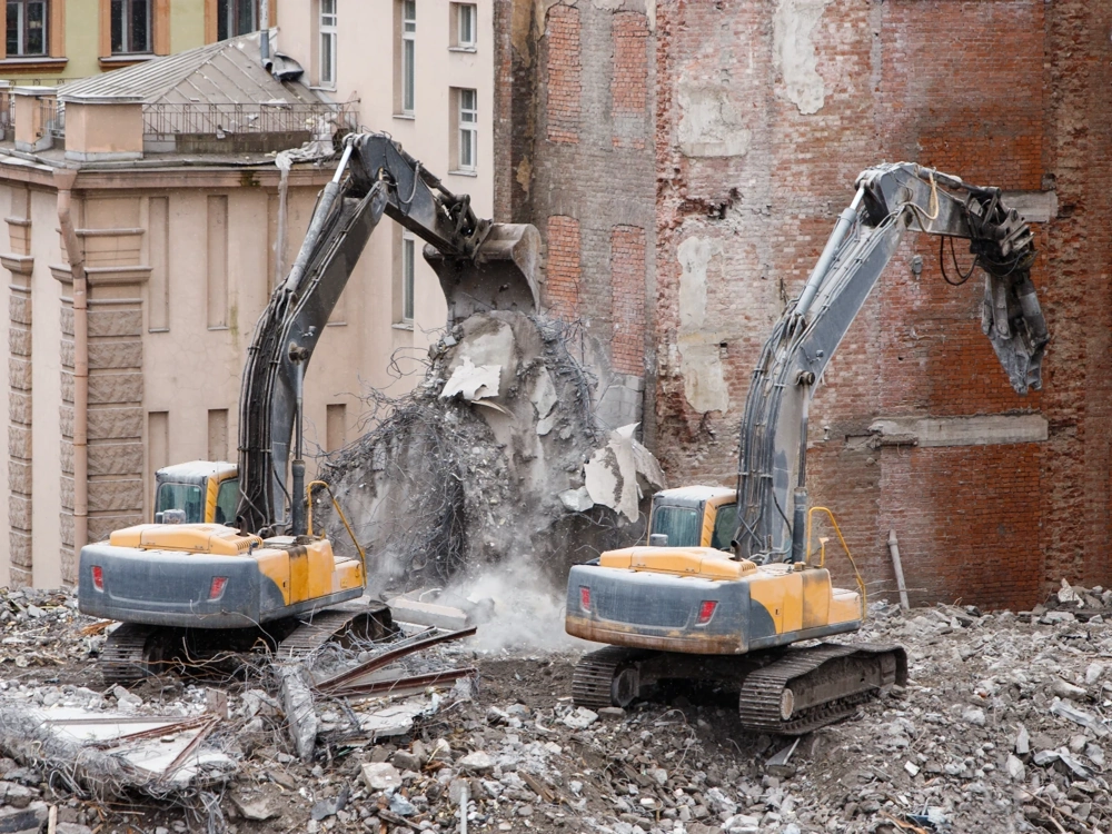 Dwie koparki kruszą ceglaną ścianę w trakcie wyburzeń budynku Wyburzenia
