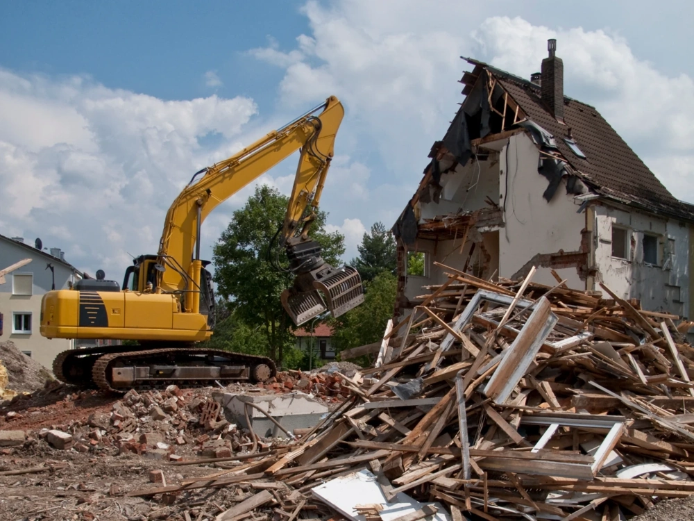 Żółta koparka rozbiera fragment domu, sterta gruzu i drewna na placu budowy – Wyburzenia
